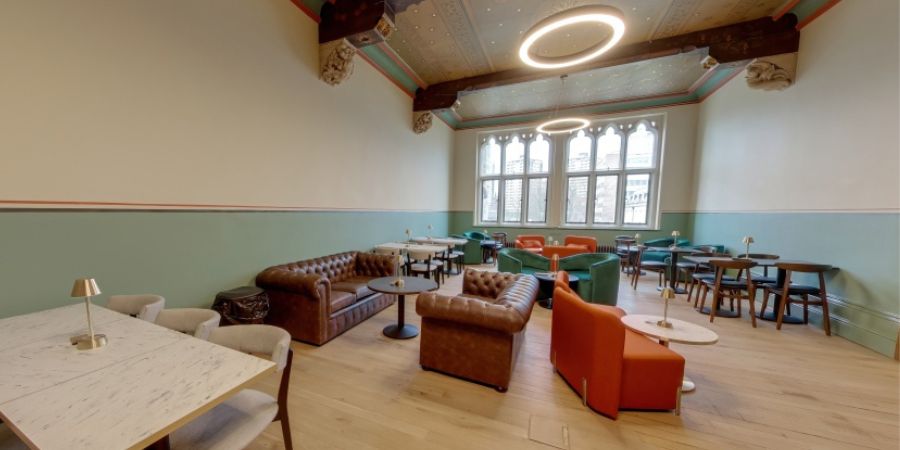 A panoramic view of a lounge area with tufted leather sofas, orange and green armchairs, and tables with lamps on a light wooden floor under a decorative ceiling.