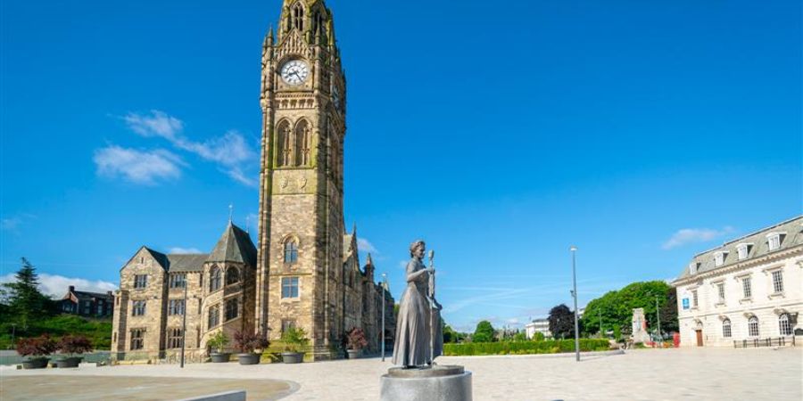 A statue of a woman in front of a large clock tower on a sunny day.
