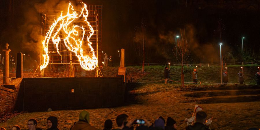 A large sculpture of a horse's head is engulfed in flames at night. A crowd watches the spectacle, some holding torches, in a park-like setting with trees and lights.
