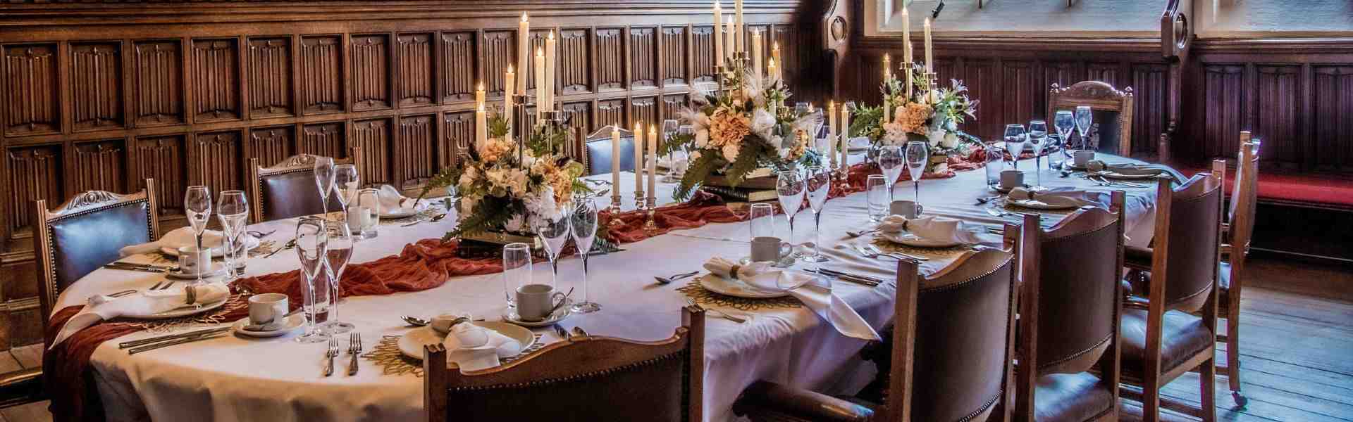 A long dining table set with white linens, floral arrangements, and lit candles, surrounded by ornately carved wooden chairs in a room with wood panelling and portraits on the wall.